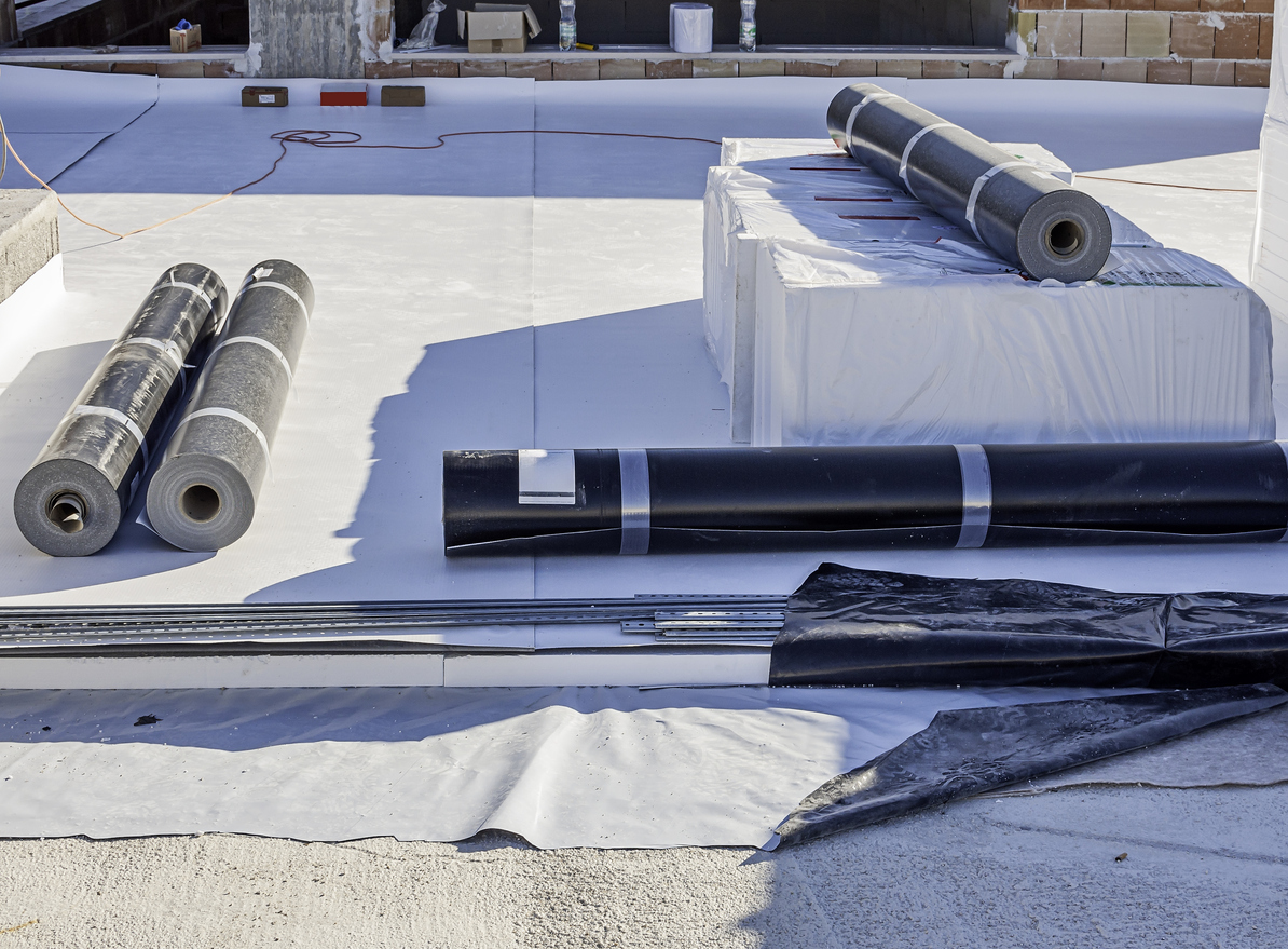 Construction site with a flat roof being waterproofed. The image shows a partially installed waterproof membrane, tools, and construction materials. The scene highlights the roofing process.